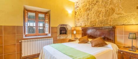 A bedroom with a stone wall, a window with shutters, a radiator, a bed with pillows, and a nightstand with a lamp.