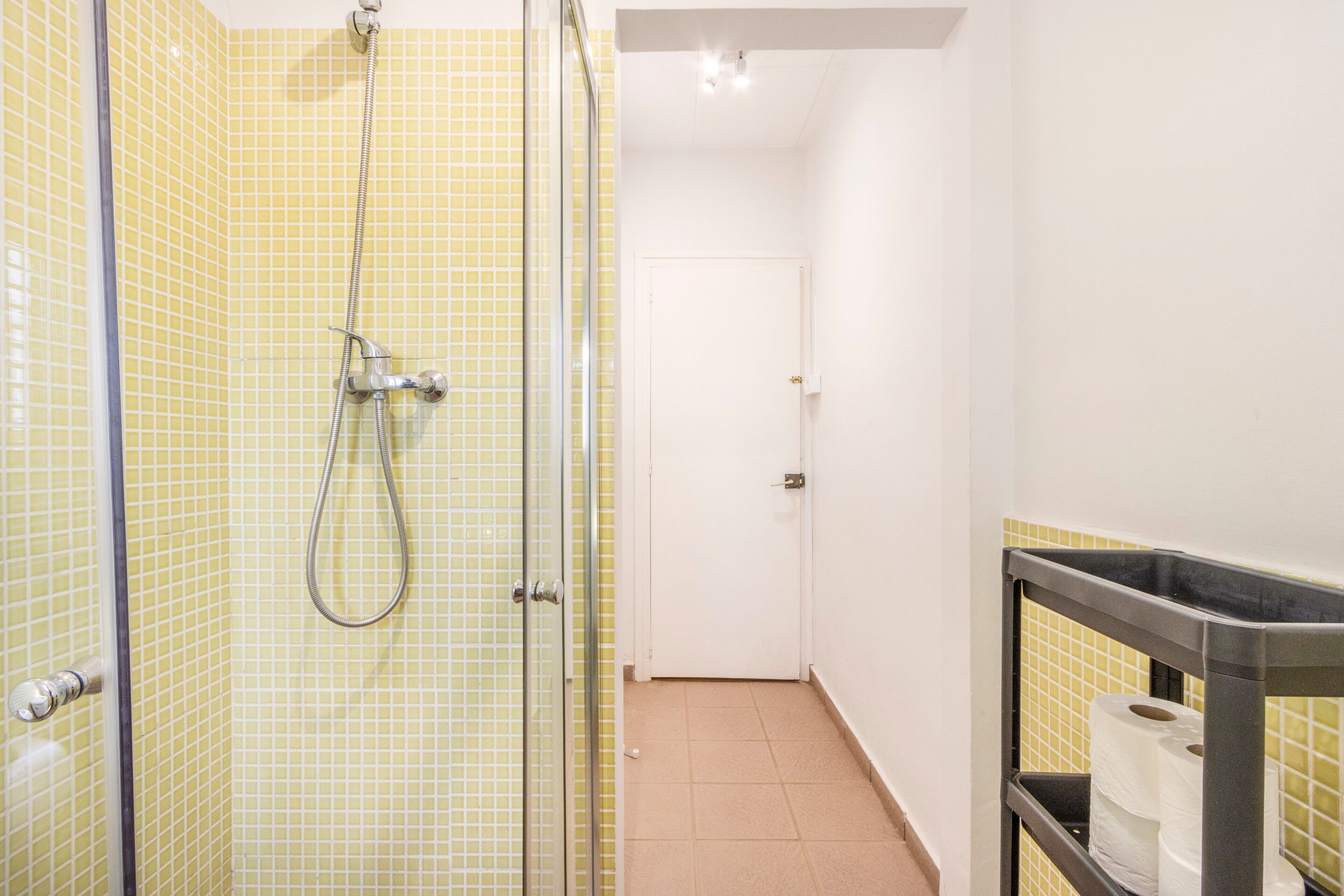 Un baño con paredes de azulejos amarillos, cabina de ducha de vidrio, una puerta blanca y un estante con papel higiénico.