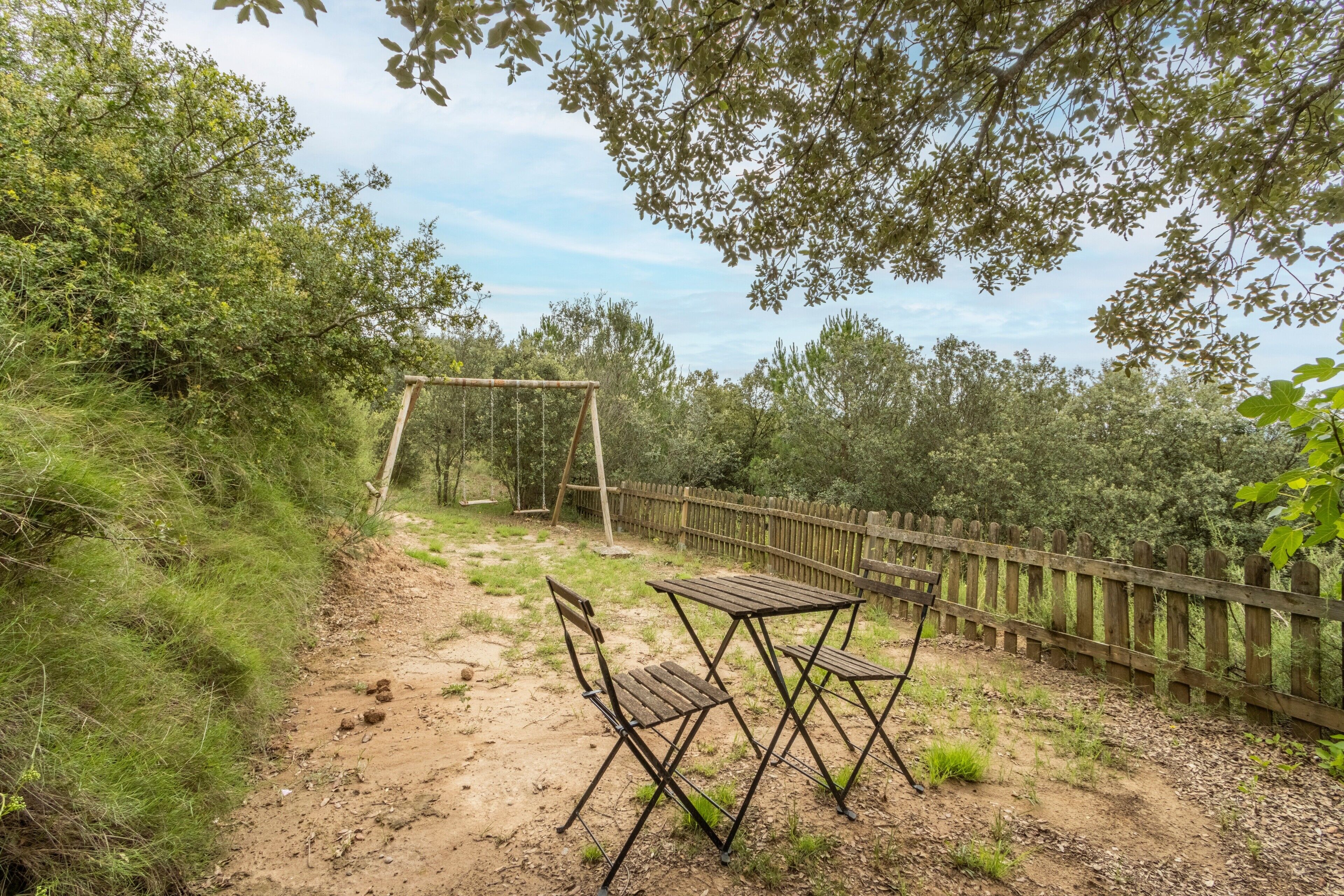 Een onverhard pad met een houten schommel en een metalen tafel met stoelen.