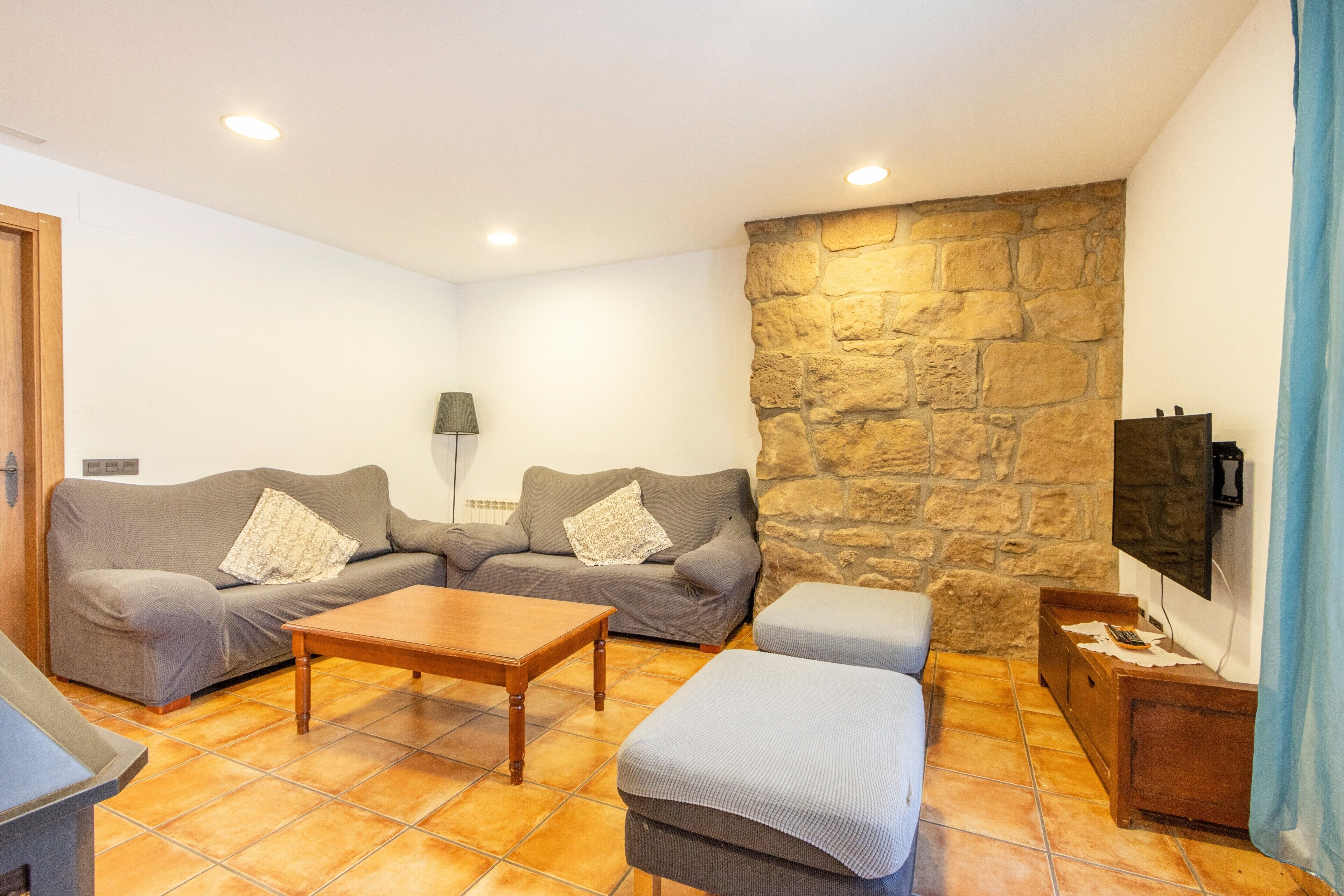 Un salon avec un mur en pierre, deux canapés gris, une table basse en bois et un téléviseur fixé au mur.