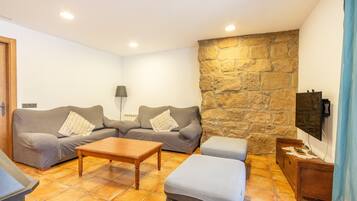 Un salon avec un mur en pierre, deux canapés gris, une table basse en bois et un téléviseur fixé au mur.