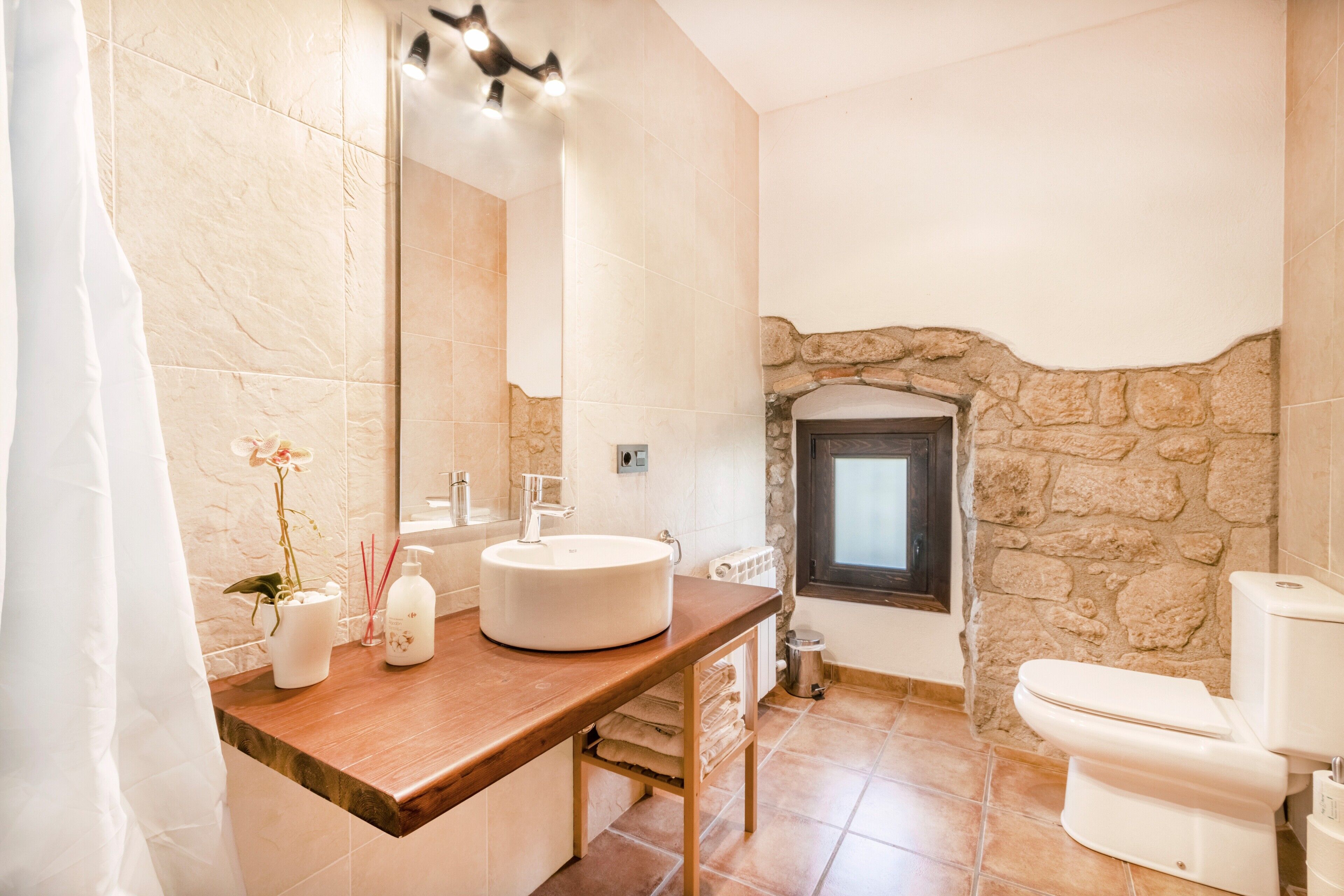 Een badkamer met een stenen muur, een witte wastafel, een houten wastafelmeubel, een spiegel, een toilet en een raam.