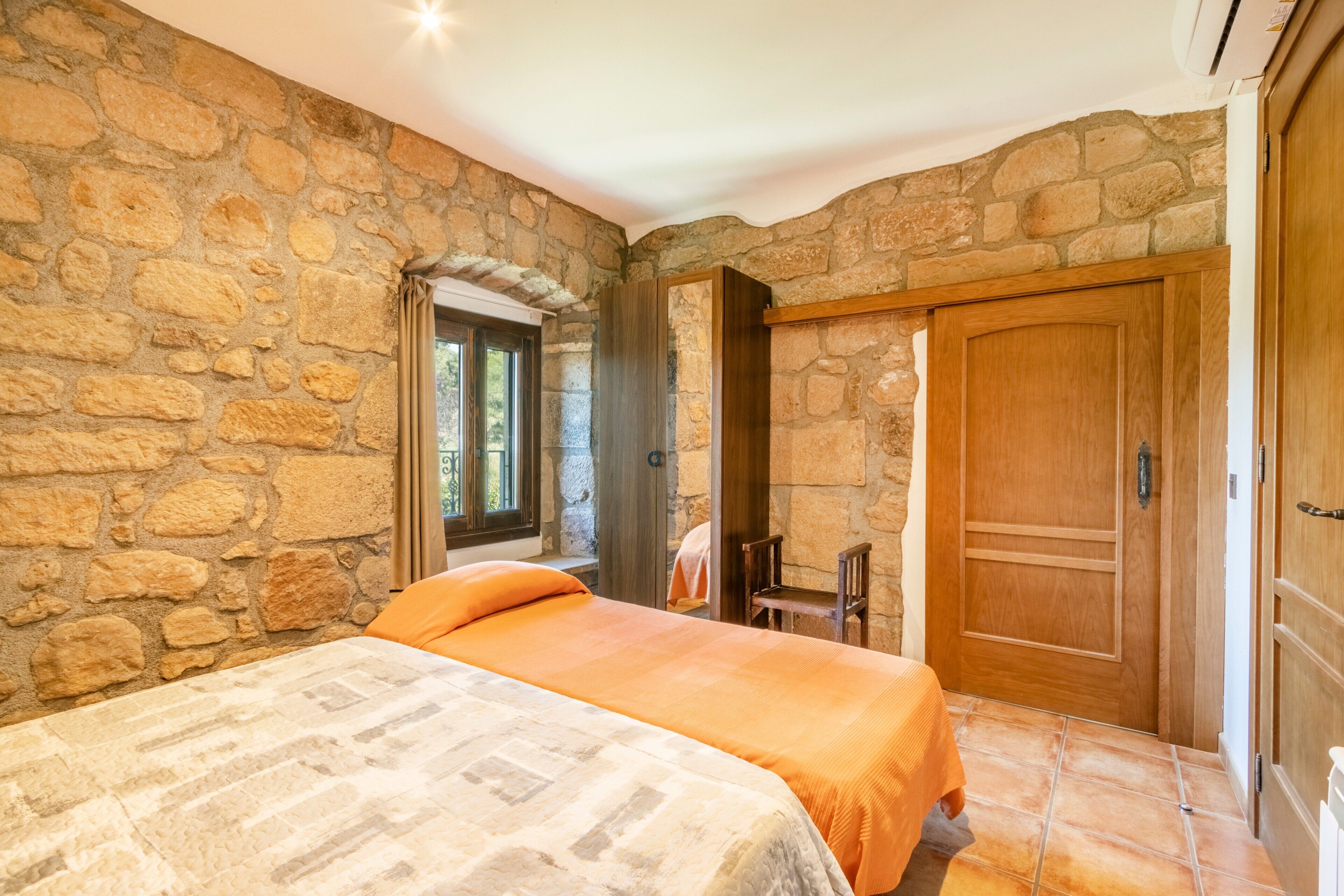 A bedroom with stone walls, a bed with an orange bedspread, a wooden door, and a window with curtains.
