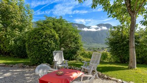 Une table recouverte d’une nappe rouge, entourée de quatre chaises blanches, elle-même bordée de gravier et d’une bordure en pierre, avec un paysage montagneux en arrière-plan.