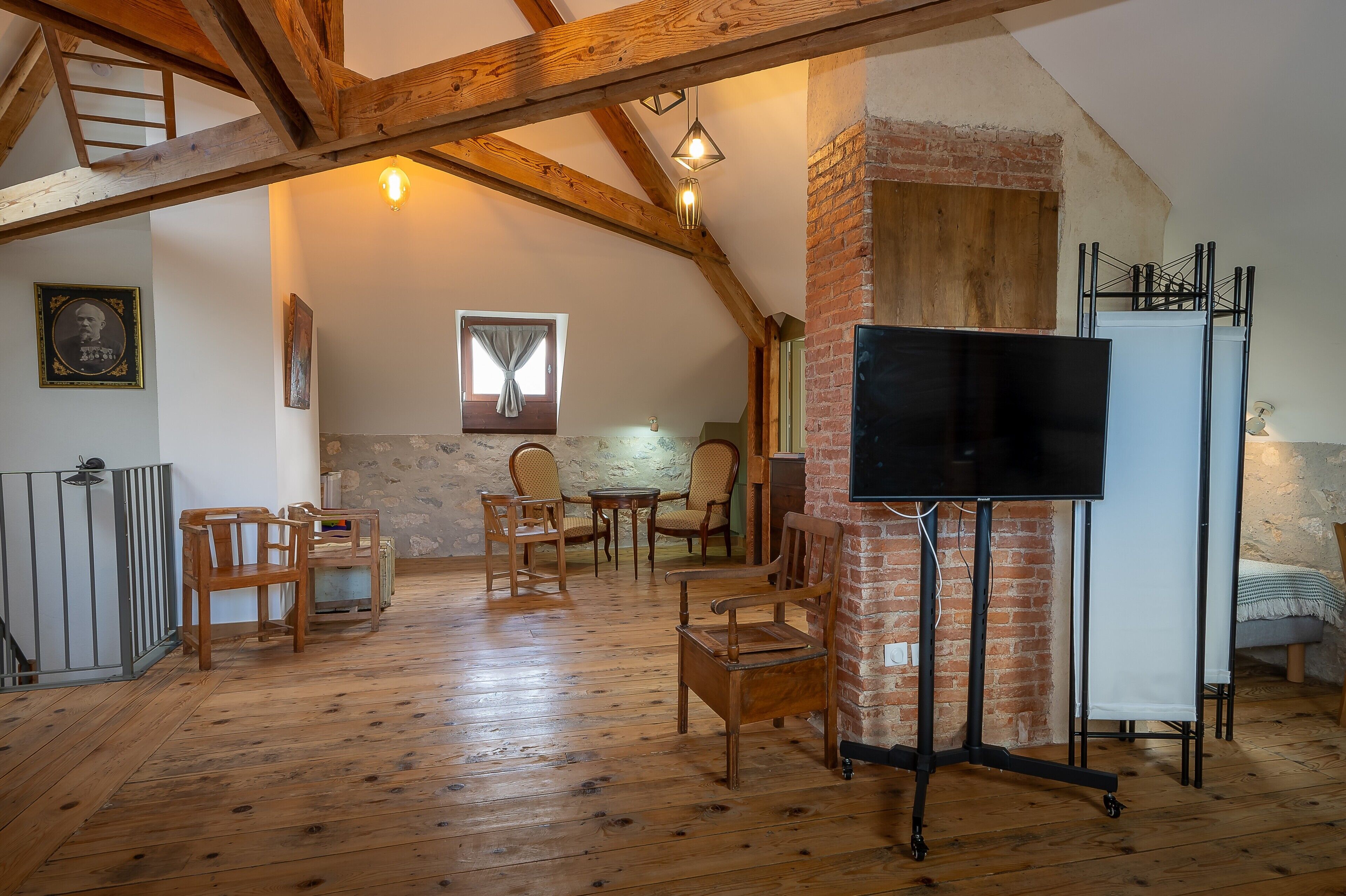 Una habitación rústica con vigas de madera a la vista, pared de ladrillo, televisor sobre un soporte y un comedor con sillas y mesa de madera.