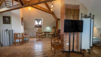 Una habitación rústica con vigas de madera a la vista, pared de ladrillo, televisor sobre un soporte y un comedor con sillas y mesa de madera.