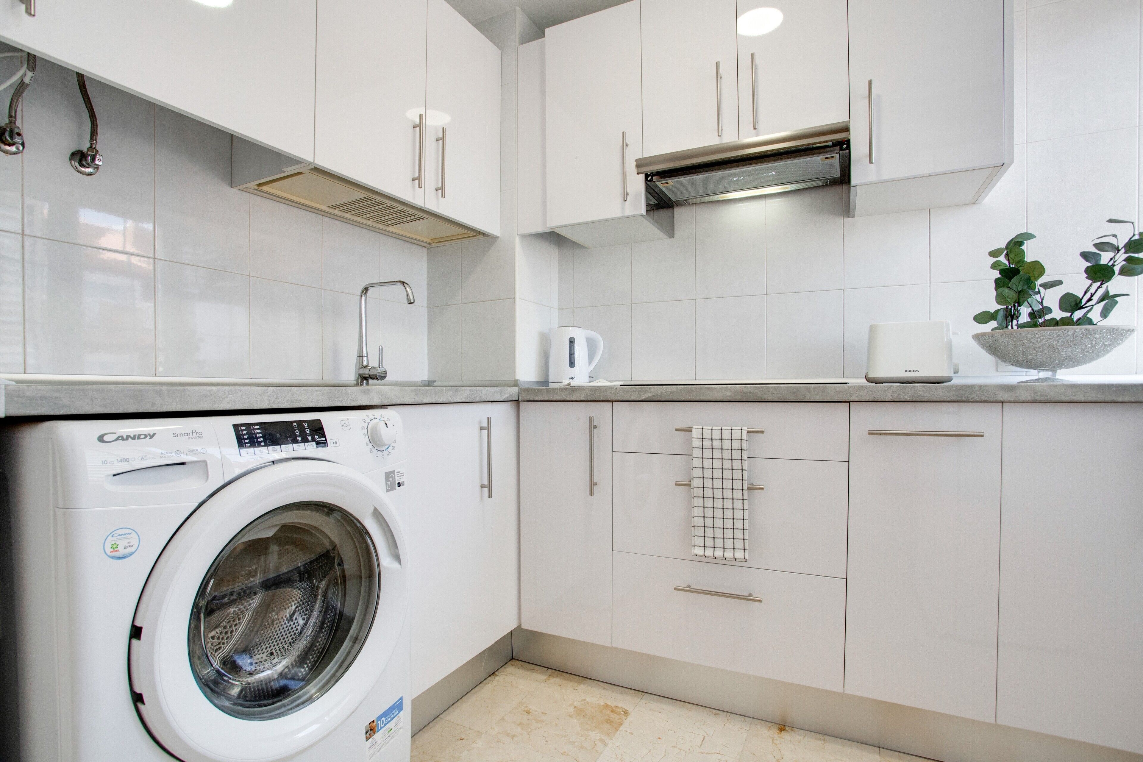 A modern kitchen with white cabinets, a built-in washing machine, a coffee maker, and a plant.