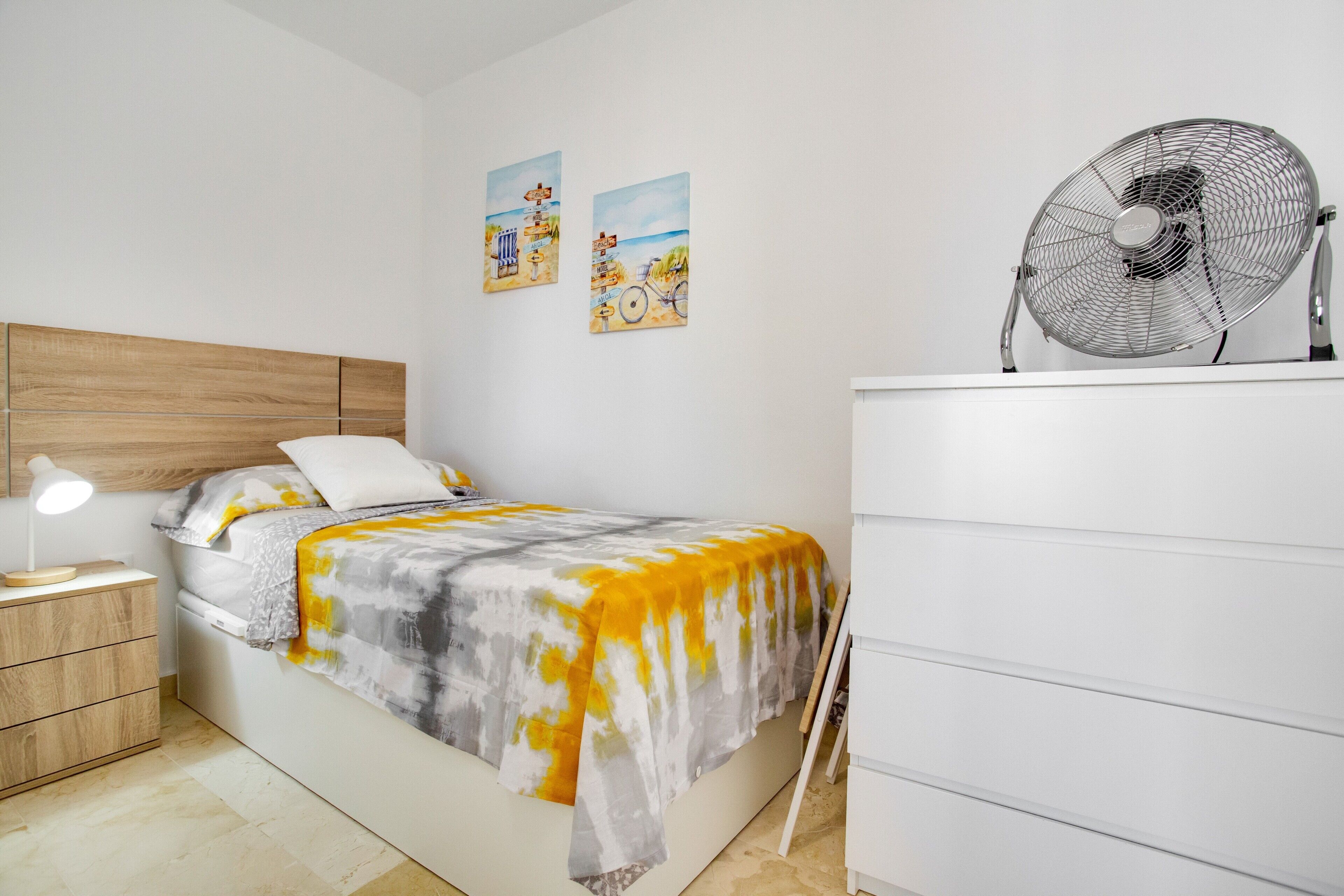 Een slaapkamer met een bed, nachtkastje, lamp, ventilator, kaptafel en twee decoraties aan de muur.