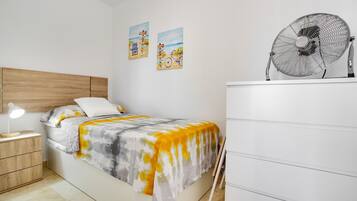 Een slaapkamer met een bed, nachtkastje, lamp, ventilator, kaptafel en twee decoraties aan de muur.
