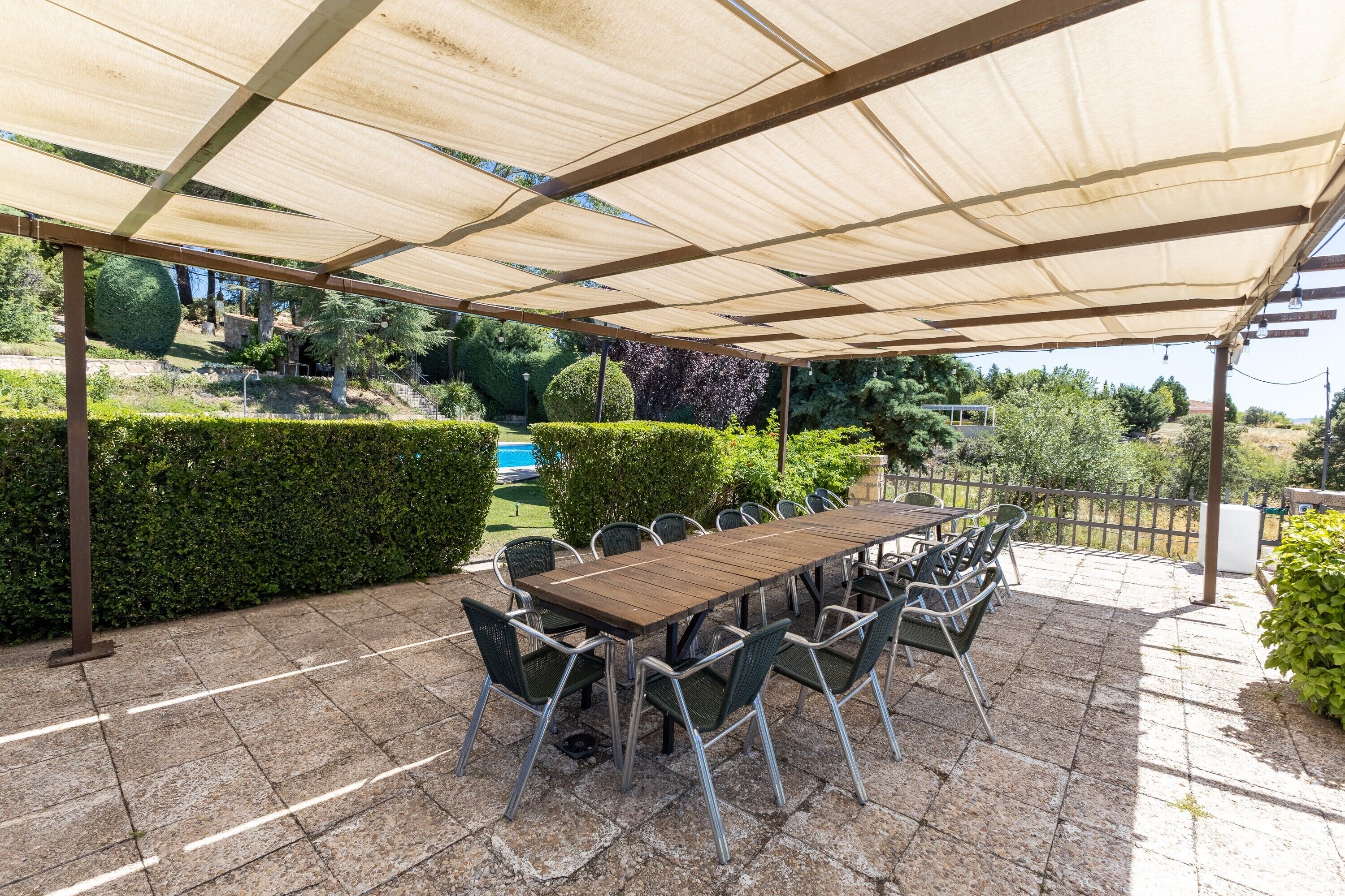Een buitenterras met een houten tafel en stoelen, een pergola erboven en een zwembad met groen in de achtergrond.