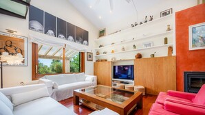Un salon moderne avec une table basse en verre, des canapés blancs et rouges, un téléviseur, des étagères en bois ornées d’objets décoratifs et une cheminée.