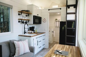 Une cuisine compacte avec des meubles blancs, un plan de travail en bois et un micro-ondes noir. Une fenêtre laisse entrer la lumière naturelle et offre une vue sur les arbres à l’extérieur.