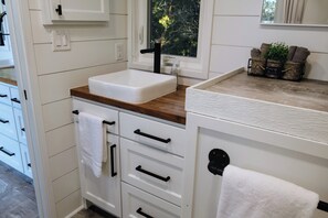 Une salle de bain moderne avec un lavabo blanc, un plan de toilette en bois et une fenêtre donnant sur les arbres.
