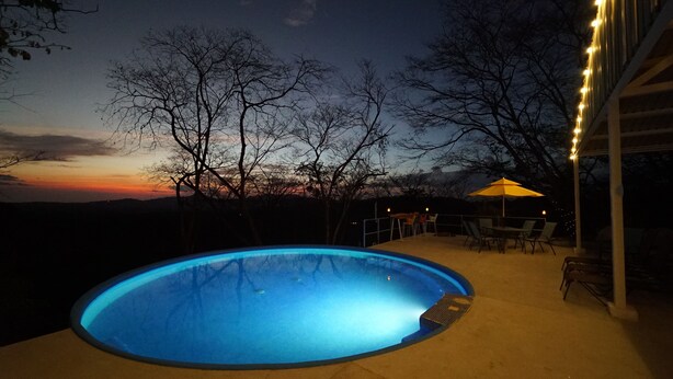 Ein Außenpool im Abenddämmerung mit Sonnenuntergangsblick, umgeben von Bäumen und einer Terrasse mit Stühlen und Sonnenschirm.