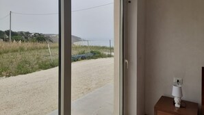 Una habitación con un amplio ventanal corredera que da a una vista de la playa, una mesita de noche con lámpara y una pared lisa.