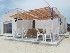 Zona d'estar a l'aire lliure coberta amb cadires de plàstic i una taula, un edifici amb una finestra gran i un sòl sorrenc.
