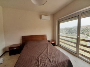 Un dormitorio con cama, mesita de noche, cajón, ventana con vistas al exterior y un equipo de aire acondicionado.