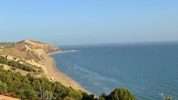 Un paysage côtier avec une plage, une colline surmontée d’une construction et un ciel dégagé.