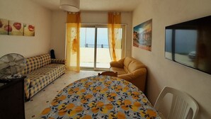 A room with a sofa, a bed with a floral bedspread, a fan, a chair, a painting, and a view through a window. - Room 'Vista Montagna 2' with Shared Terrace, Wi-Fi and Air Conditioning (Siculiana)