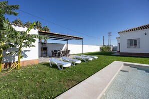 A backyard with a swimming pool, lounge chairs, a patio with tables and chairs, and a covered area.