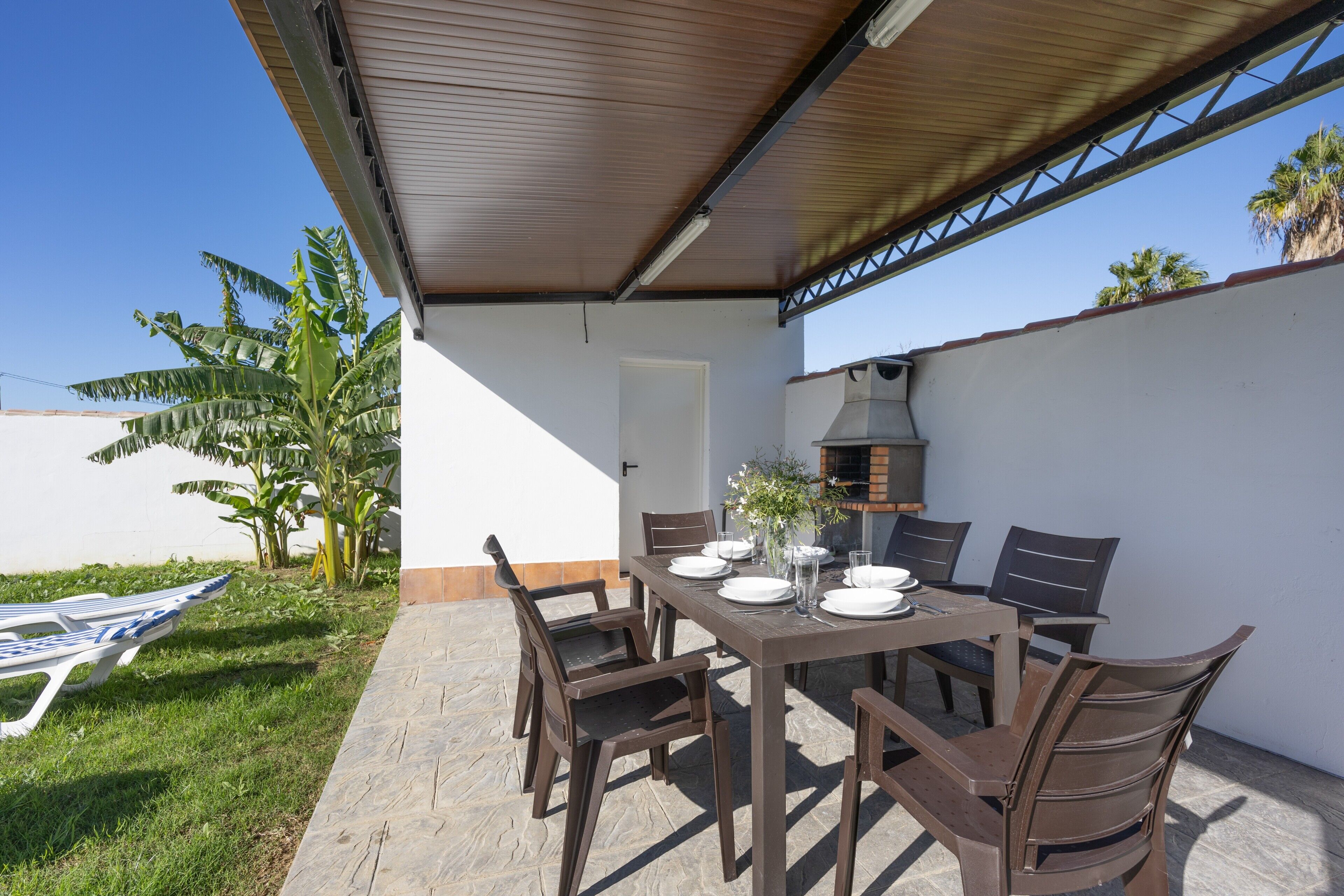 An outdoor patio with a table set for dining, surrounded by chairs, a grill, and a covered area with a door.