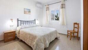 A bedroom with a bed, bedside table, chair, window with curtains, and an air conditioning unit.