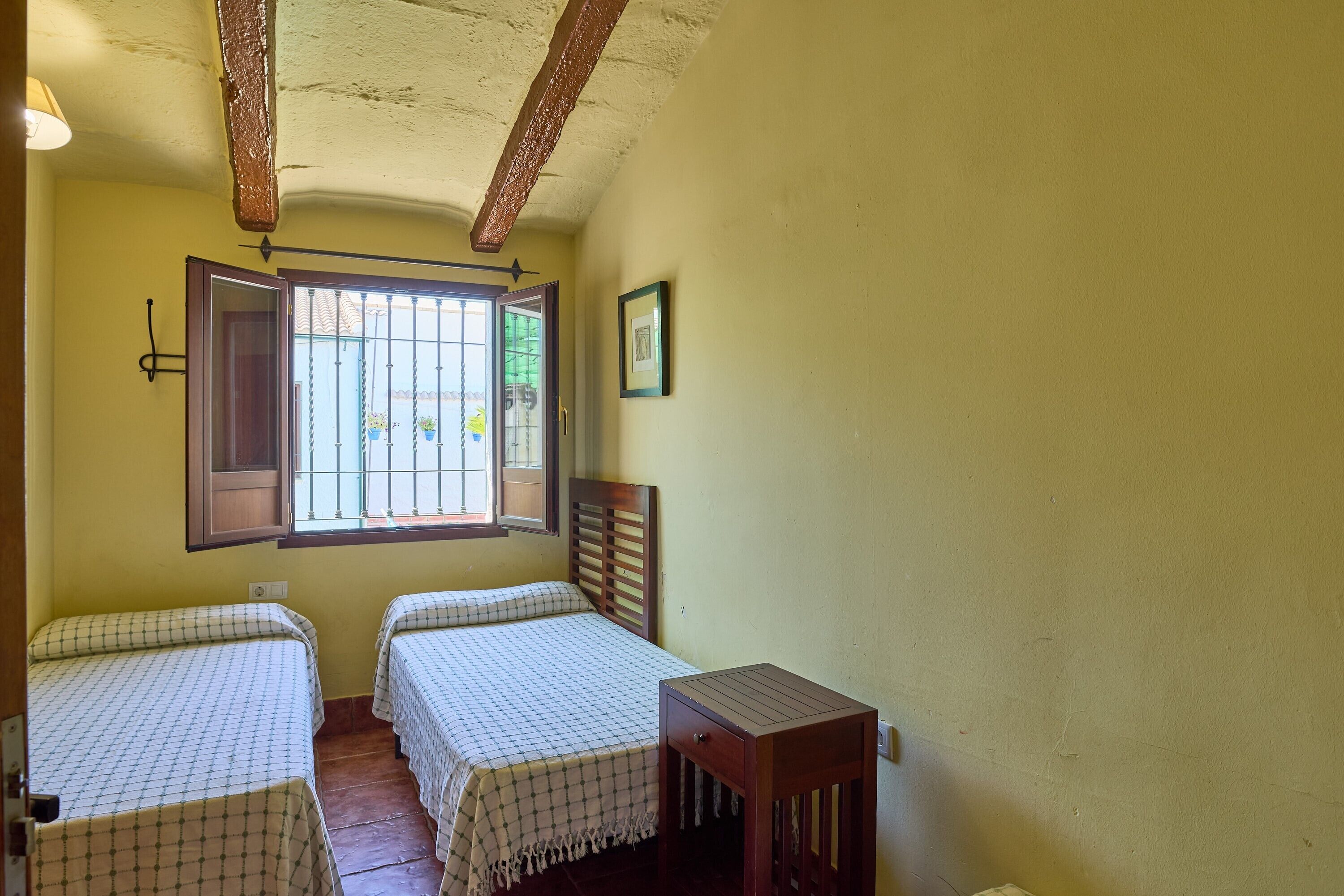 Una habitación con dos camas, una mesita, una ventana con barrotes y un cuadro en la pared.