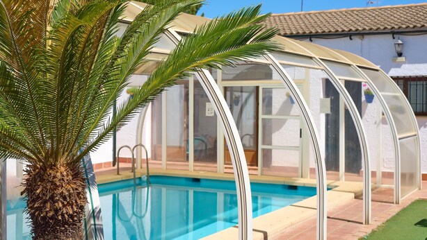Una piscina con cerramiento de vidrio, una palmera y un edificio con techo de tejas.