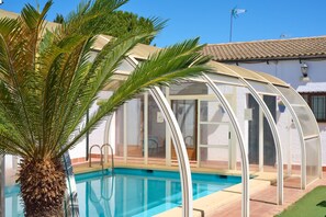 A swimming pool with a glass enclosure, a palm tree, and a building with a tiled roof. - Rivero Rural Country House - Private and Indoor Pool, 10 Bedrooms, 22 Guests (Rivero de Posadas)