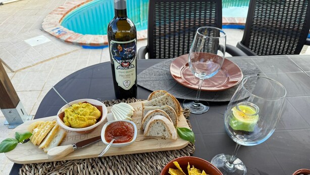 A table set with a bottle of wine, a glass, a bowl of chips, a bowl of dip, slices of bread, and a plate of cheese.