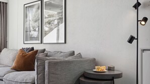 Un salon moderne avec un canapé gris, un coussin marron et une petite table ronde sur laquelle se trouve une assiette de nourriture. Deux tableaux encadrés ornent le mur, et une lampe sur pied est placée dans un coin.