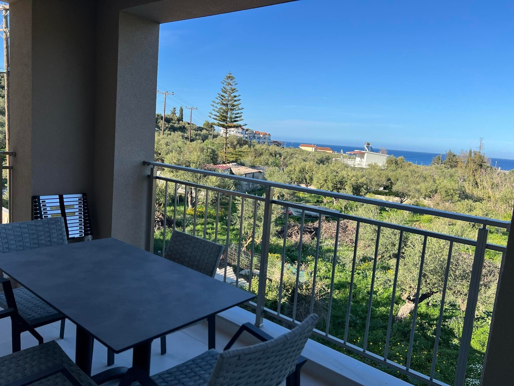 Ein Balkon mit Tisch und Stühlen, der auf eine Küstenlandschaft mit Bäumen und Gebäuden blickt.