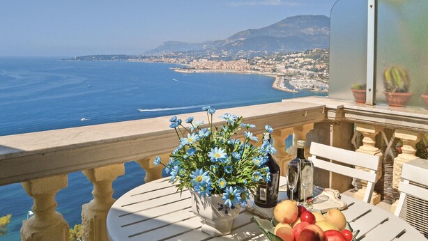 Un balcon avec une table ornée d’un vase rempli de fleurs bleues, d’une bouteille et d’un verre de vin, d’où l’on peut admirer une vue sur le paysage urbain de la côte.