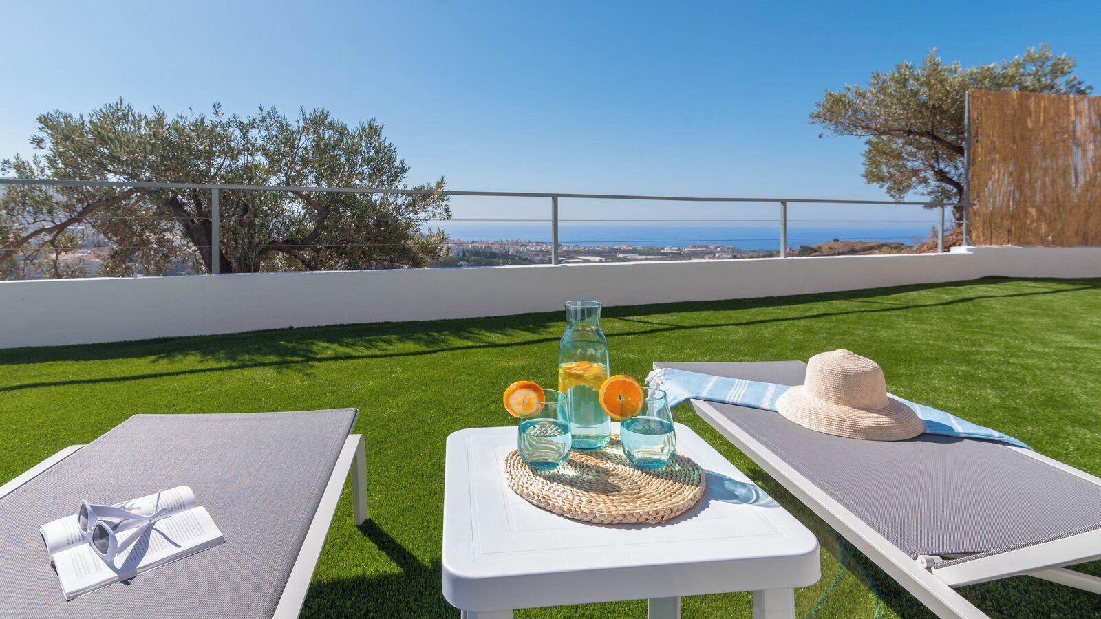 A terrace with two lounge chairs, a table with drinks and a book, and a view of the sea.