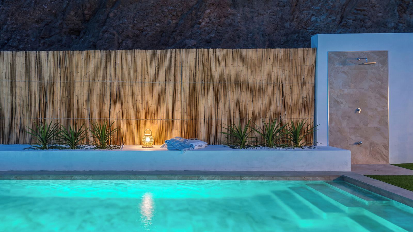 Ein Schwimmbecken mit Bambuswand im Hintergrund, ein kleiner Tisch mit einer Lampe und eine Tür rechts.