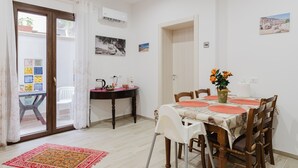A dining area with a table set for four, a chair, a small table with a vase of flowers, and a view of a building through a window. - Holiday Home 'Iris Castelvetrano' with Wi-Fi and Air Conditioning (Castelvetrano)