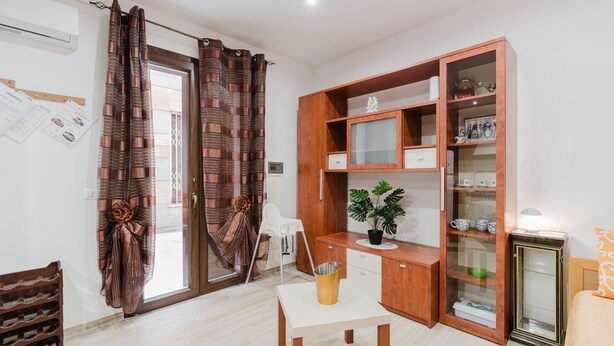 Una habitación con un escritorio de madera, una silla, una mesita con una planta, un aparador con puertas de cristal y un zapatero.
