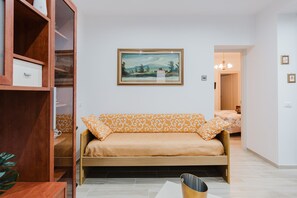 A room with a sofa bed, a wooden cabinet, a framed landscape painting, and a hallway leading to another room.