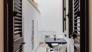 Un pasillo estrecho con una mesa, sillas, una planta en maceta y un estante metálico con objetos.