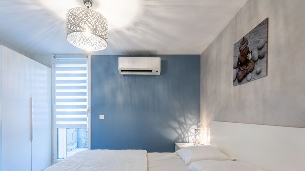 Un dormitorio con cama, ropa de cama blanca, un equipo de aire acondicionado, una obra de arte en la pared y una lámpara en el techo.