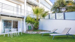 Un edificio moderno con un balcón, un pequeño jardín con una palmera y dos sillones de relax.