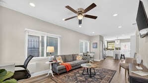 A spacious living area with a grey sofa, a coffee table, and a ceiling fan. There is a kitchen in the background with white cabinets and a dining area with a table and chairs.