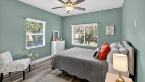A bedroom with a bed, a chair, a dresser, a ceiling fan, and two windows.