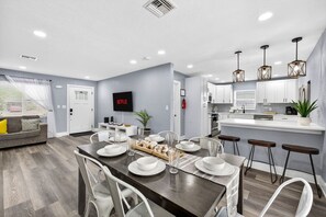 Une cuisine moderne avec des meubles blancs, un coin repas doté d’une table en bois pour quatre personnes, et un salon avec un canapé et un téléviseur fixé au mur.