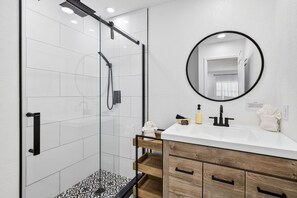 A modern bathroom with a glass-enclosed shower, a white sink with a black faucet, a wooden vanity, and a round mirror. - Family Pool Home | Game Room + Fire Pit | Largo (Largo)