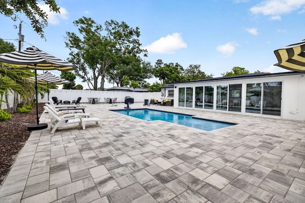 Piscina com espreguiçadeiras e guarda-sóis, rodeada por um pátio pavimentado e um edifício branco com grandes janelas.