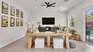 A modern dining area with a wooden table, two white cushioned benches, a ceiling fan, and a large TV mounted on the wall. - Heated Pool & Spa, Golf Simulator, Billiards (Scottsdale)