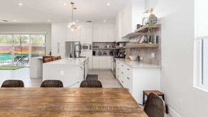 A modern kitchen with white cabinetry, a central island, and a dining area with a wooden table. - Heated Pool & Spa, Golf Simulator, Billiards (Scottsdale)