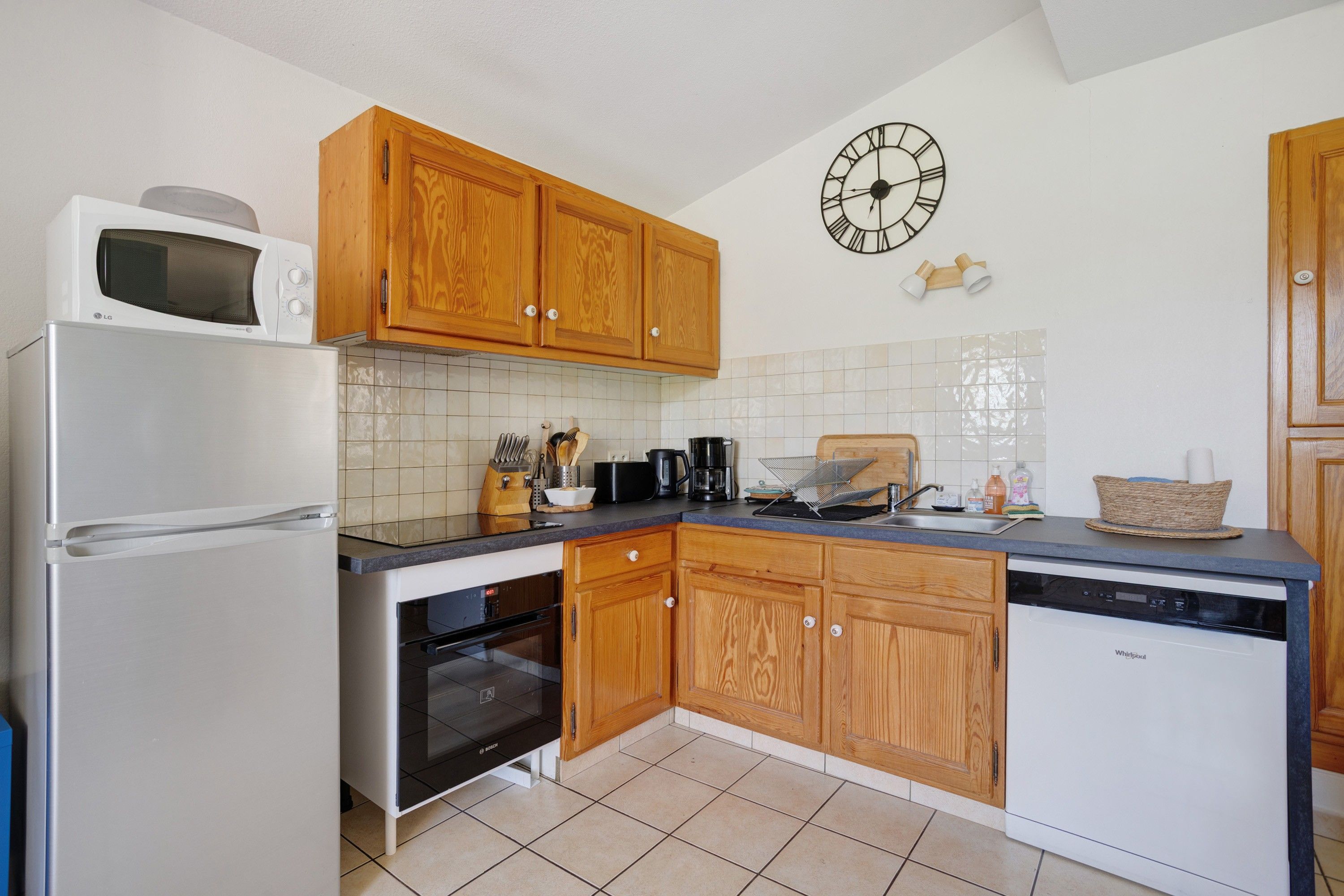 A compact kitchen with wooden cabinets, a microwave, a Bosch dishwasher, and a clock on the wall.