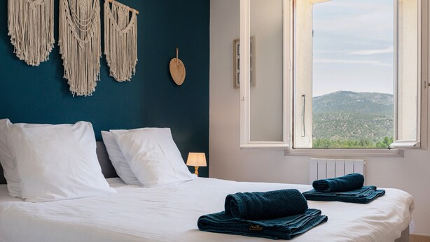 A bed with white linens, a macrame wall hanging, a view through a window, and a folded towel on the bed.